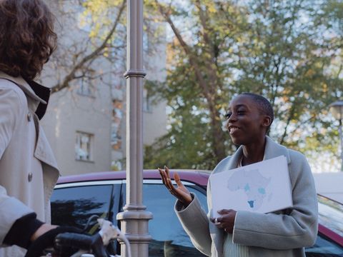 Bildvergrößerung: Eine Frau steht auf einem Gehweg und redet in die Richtung einer anderen Person. Die Frau hält eine Karte von Afrika mit markierten Bereichen in der Hand.