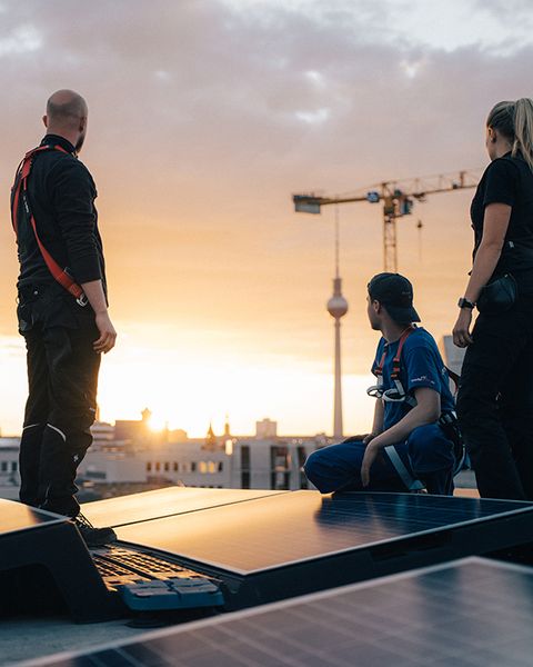 Drei Personen bei Sonnenaufgang auf dem Dach, im Vordergrund Solarpanel, im Hintergrund die Stadt Berlin.