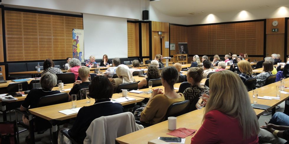 Viele Menschen sitzen in Tischreihen in einem Saal und schauen zu drei Frauen dir vorne an Mikrofonen sitzen.