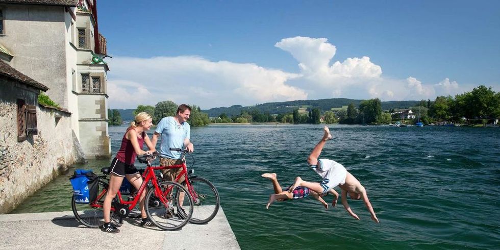 Zwei Radfahrer am Bodensee schauen zu wie zwei Menschen von einer Mauer in den See springen