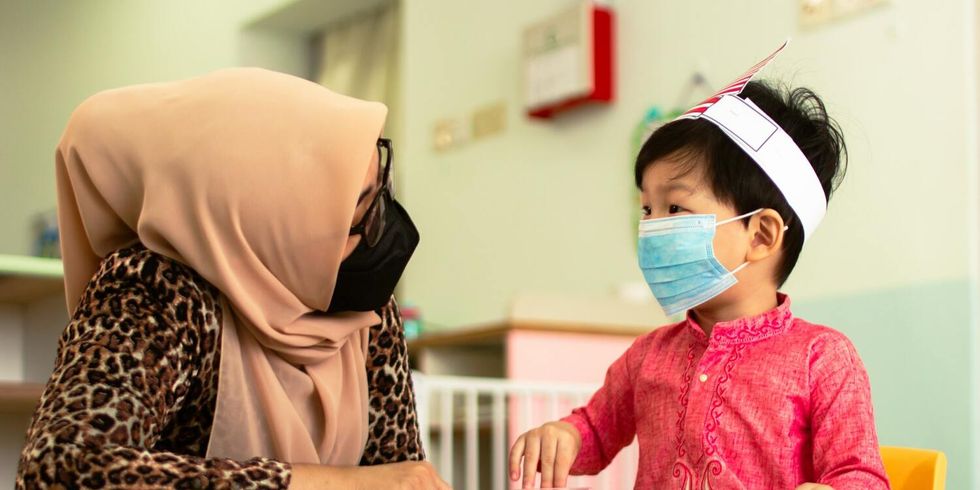 Erzieherin einer Kindertagesstätte und ein Kind sitzen am Tisch und unterhalten sich. Beide tagen eine medizinische Maske, um sich vor dem Coronavirus zu schützen.