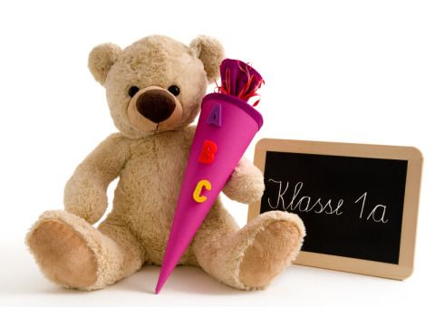 Teddybär mit einer Schultüte mit der Aufschrift "ABC" und einer Tafel auf der "Klasse 1a" steht