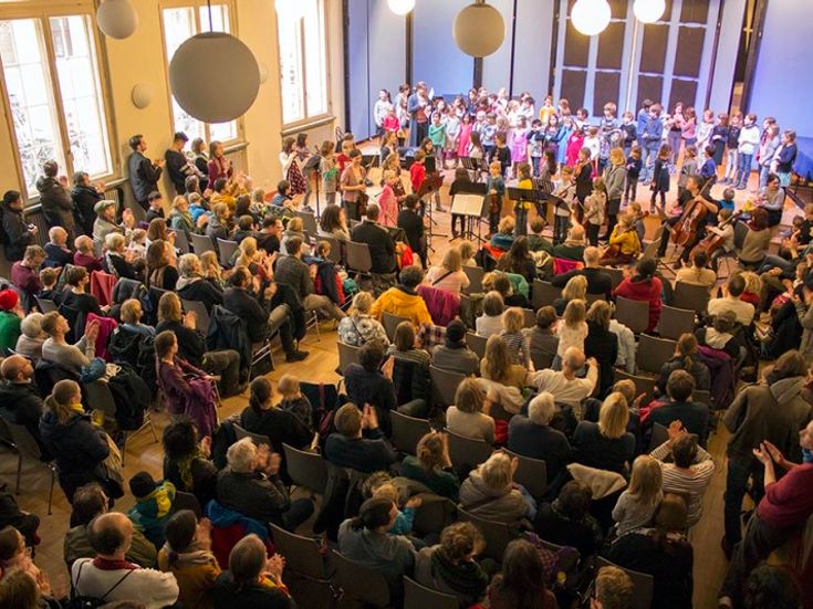 Voller Konzertsaal mit Bühne, darauf singende und musizierende Kinder