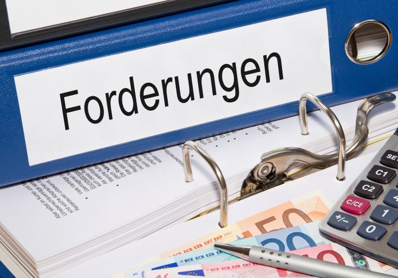 Auf einem geöffneten Aktenordner liegen ein Taschenrechner, ein Stift, mehrere Geldscheine und weitere Ordner mit der Aufschrift 'Forderungen'