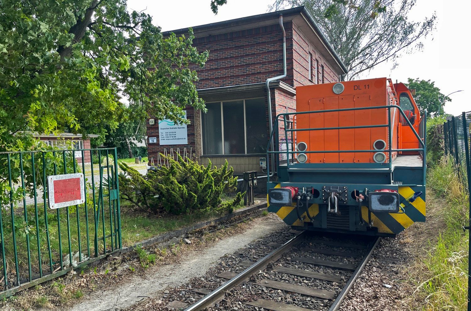 Orange Lok auf dem Gleis umrahmt von Bäumen, im Hintergrund ein Backsteingebäude.