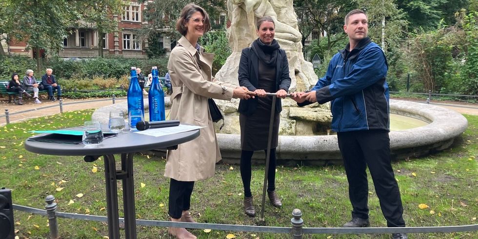 Zwei Frauen und ein Mann halten zusammen einen Drehschieber und stehen auf einer Grünfläche vor einem nicht aktiven Brunnen.