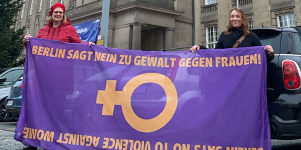 Zwei Frauen stehen vor dem rathaus Schöneberg und halten eine Lila/orangene fahne hoch mit der Aufschrift: Berlin sagt Nein zu Gewalt an Frauen.