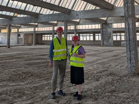 Bildvergrößerung: Eine Frau und ein Mann, beide mit Warnweste und Helm gekleidet, stehen in einer großen leeren Halle.