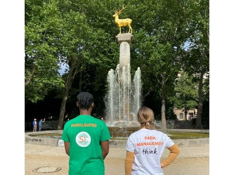 Bildvergrößerung: Ein Mann und eine Frau stehen mit dem Rücken zur Kamera in einem Park und schauen zu einem Springbrunnen mit der Statue eines goldenen Hirschs. Der Mann trägt ein grünes T-Shirt mit der Aufschrift "Parkläufer" und die Frau trägt ein weißes T-Shirt mit der Aufschrift "Parkmanagement".