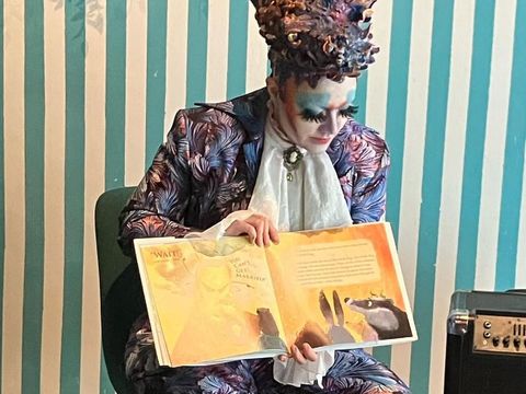 Bildvergrößerung: Eine bunt gekleidete Person mit starkem Makeup und bunten Haaren sitzt auf einem Stuhl und schaut auf ein Bilderbuch, was dem Zuschauer zugewandt ist.