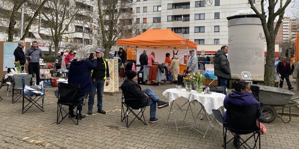 Auf einem Platz stehen und sitzen Menschen. In der Mitte ist ein orangenes Zelt.