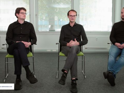 Drei Männer sitzen auf Stühlen in einem leeren Raum, einer spricht