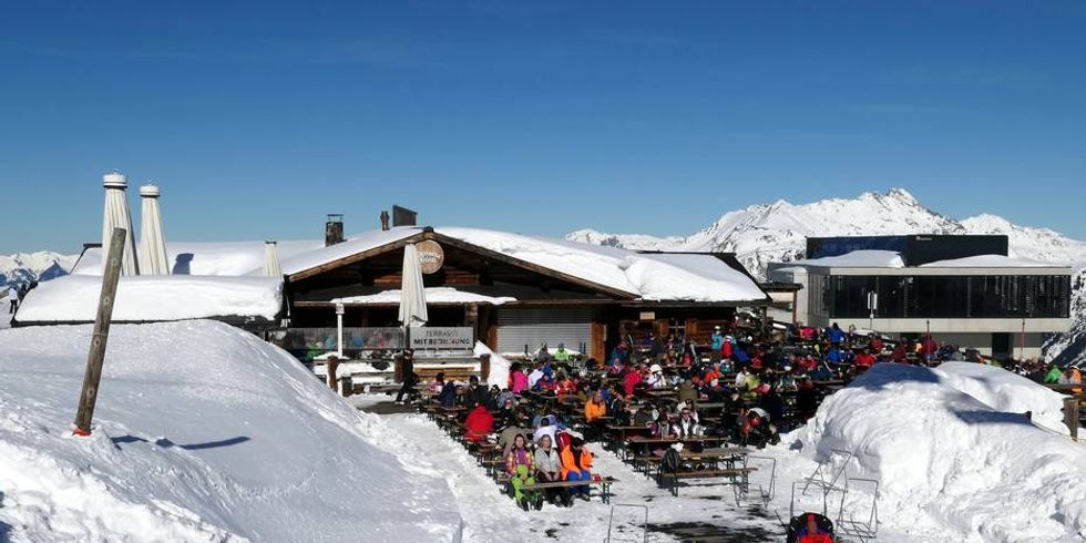 verschneite Berglandschaft mit Baude im Vordergrund davor sitzen Personen welche essen und trinken