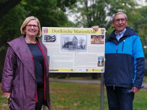 Bildvergrößerung: eine Frau und ein Mann stehen vor einem Schild mit viel Text und der Überschrift Dorfkirche Marienfelde