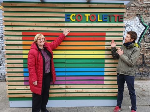 Zwei Personen stehen vor einem kleinen Gebäude mit der Aufschrift „Eco Toilette“