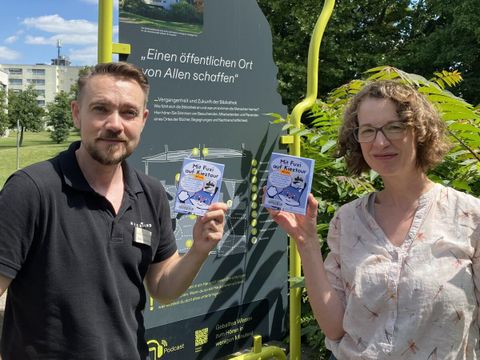 Bildvergrößerung: Zwei Personen stehen vor einer Grünfläche und einer Infotafel und halten eine Broschüre mit dem Titel Mit Fuxi auf Kieztour in der Hand.