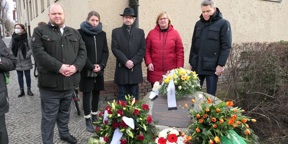 Fünf Menschen stehen zusammen hinter einem Gedenkstein. Auf diesem Stein sind mehrere Blumensträuße abgelegt.