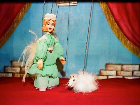 Puppentheater-Szene: Frau mit weißen Federn auf dem Rücken begegnet ein weißes, fluffiges Tier.