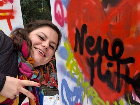 Bildvergrößerung: Eine Frau schaut seitlich ins Bild und zeigt mit einer Hand das Peace-Zeichen. Neben ihr steht eine mit Graffiti besprayte Aufstellwand, in der Mitte ein rotes Herz mit dem Schriftzug "Neue Mitte".
