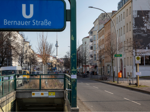 eine Nahaufnahme des Eingangs zum U-Bahnhof Bernauer Straße. Im Hintergrund sind macht im verlauf der Straße den Fernsehturm am Horizont.