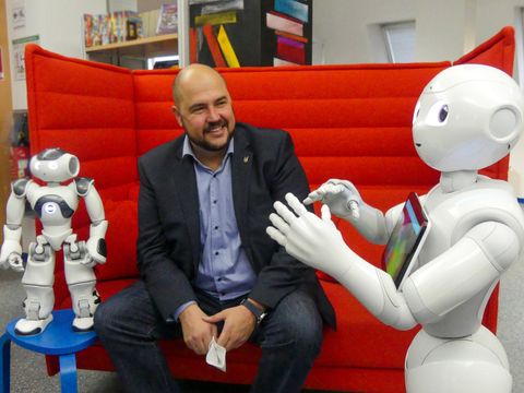 Bildvergrößerung: Bezirksstadtrat Frank Bewig auf einer roten Couch sitzend zwischen den beiden humanoiden Robotern