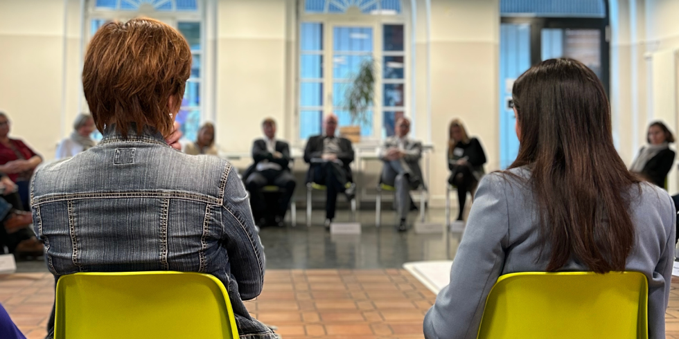 Das Bild zeigt zwei Frauen von hinten, sie sitzen auf grünen Stühlen und blicken auf eine Reihe von Menschen, die man unscharf im Hintergrund sieht. 