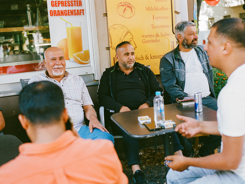 Eine Gruppe älterer Männer sitzt entspannt an einem Tisch vor einem Café in Neukölln. Das Bild fängt die Gemeinschaft und das gesellige Miteinander im Kiez ein.