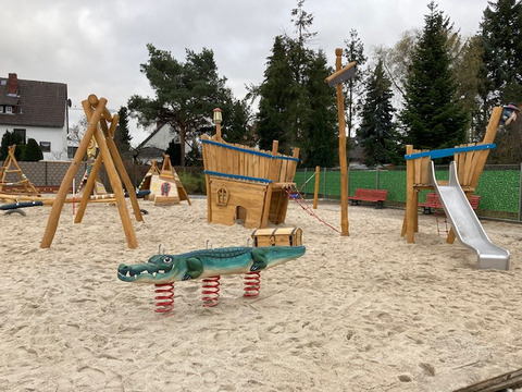 Bildvergrößerung: Ein Spielplatz mit Sandboden. Im Vordergrund sind ein Krokodil zum Schaukeln, eine Schatzkiste und eine Rutsche.