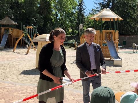 Bildvergrößerung: Eine Frau und ein Mann schneiden ein Band vor einem Spielplatz durch.