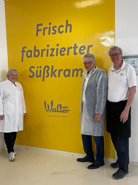 Bildvergrößerung: Drei Personen stehen vor einer Wand auf der "Frisch fabrizierter Süßkram" und das Logo der Walter Confiserie GmbH steht.
