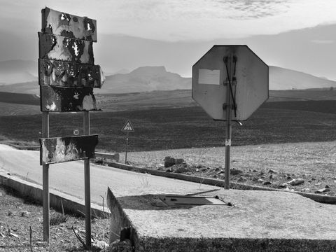 Landschaftsfotografie in schwarz-weiß, im Vordergrund eine leere Asphaltstraße mit verrosteten Schildern durch eine weite Ebene, im Hintergrund sind Berge im Dunst zu erkennen.