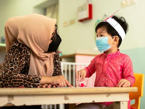 Erzieherin einer Kindertagesstätte und ein Kind sitzen am Tisch und unterhalten sich. Beide tagen eine medizinische Maske, um sich vor dem Coronavirus zu schützen.