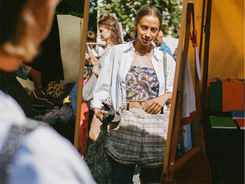 Eine Frau betrachtet sich in einem Spiegel auf einem Flohmarkt. Sie trägt ein modisches Outfit und sucht nach Secondhand-Schätzen. Die Szene vermittelt den Reiz des nachhaltigen Konsums.