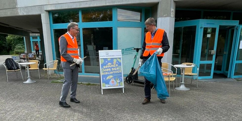 Zwei Männer mit orangenen Westen, Greifzangen und einem Müllsack stehen vor dem Eingang eines Gebäudes.