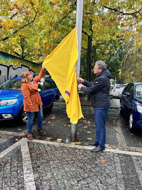 Bildvergrößerung: Eine Frau und ein Mann stehen neben einem Fahnemast und ziehen eine gelbe Fahne mit lila Kreis hoch.