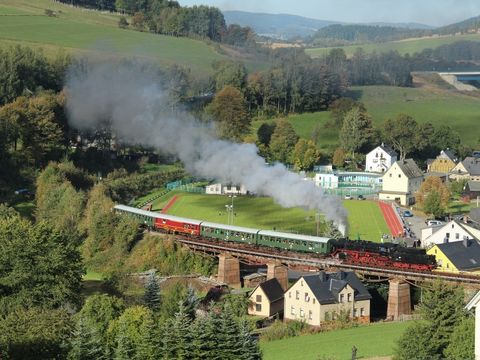 über eine Brücke fahrender Zug mit Dampflok zwischen Häusern und Wiesen