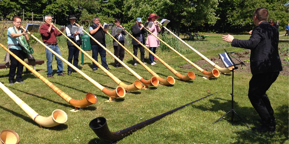 Viele Menschen spielen nebeneinander in der Natur Alphorn. Vor ihnen steht ein Dirigent.