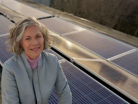 Einer lächelnde Frau mit kurzen Haaren vor Solarpanelen auf einem Dach.