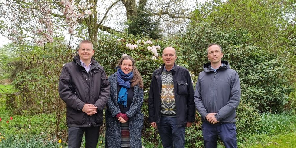 Vier Personen stehen auf einer Grünfläche. Im Hintergrund sind verschiedene Büche, Bäume, Sträucher und Blumen.