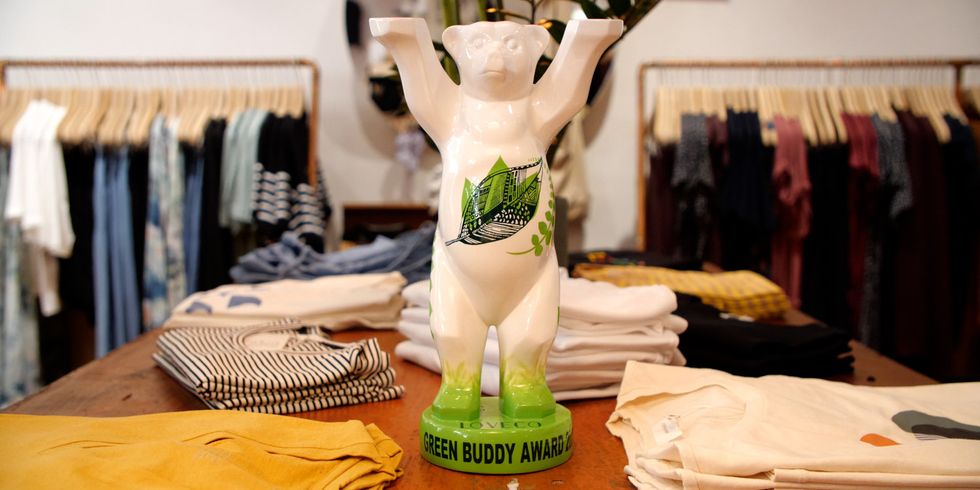 Eine Bären-Figur mit grünen Natur-Muster und dem Schriftzug "Green Buddy Award" steht auf einem Tisch umgeben von Kleidungsstücken. Im Hintergrund sieht man mehr Kleidung auf Bügeln.