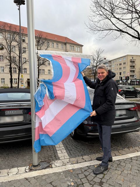 Bildvergrößerung: Ein Mann steht neben einem Fahnenmast und hält daran eine blau-rosa-weiß gestreifte Flagge hoch.