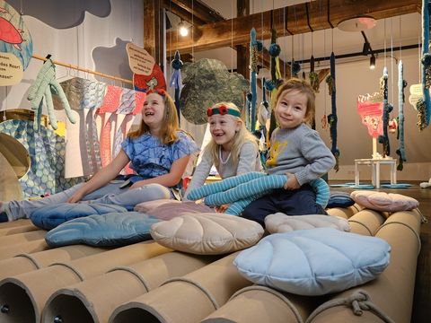 Bildvergrößerung: Drei Kinder sitzen fröhlich zwischen muschelförmigen Kissen auf einem Floß.