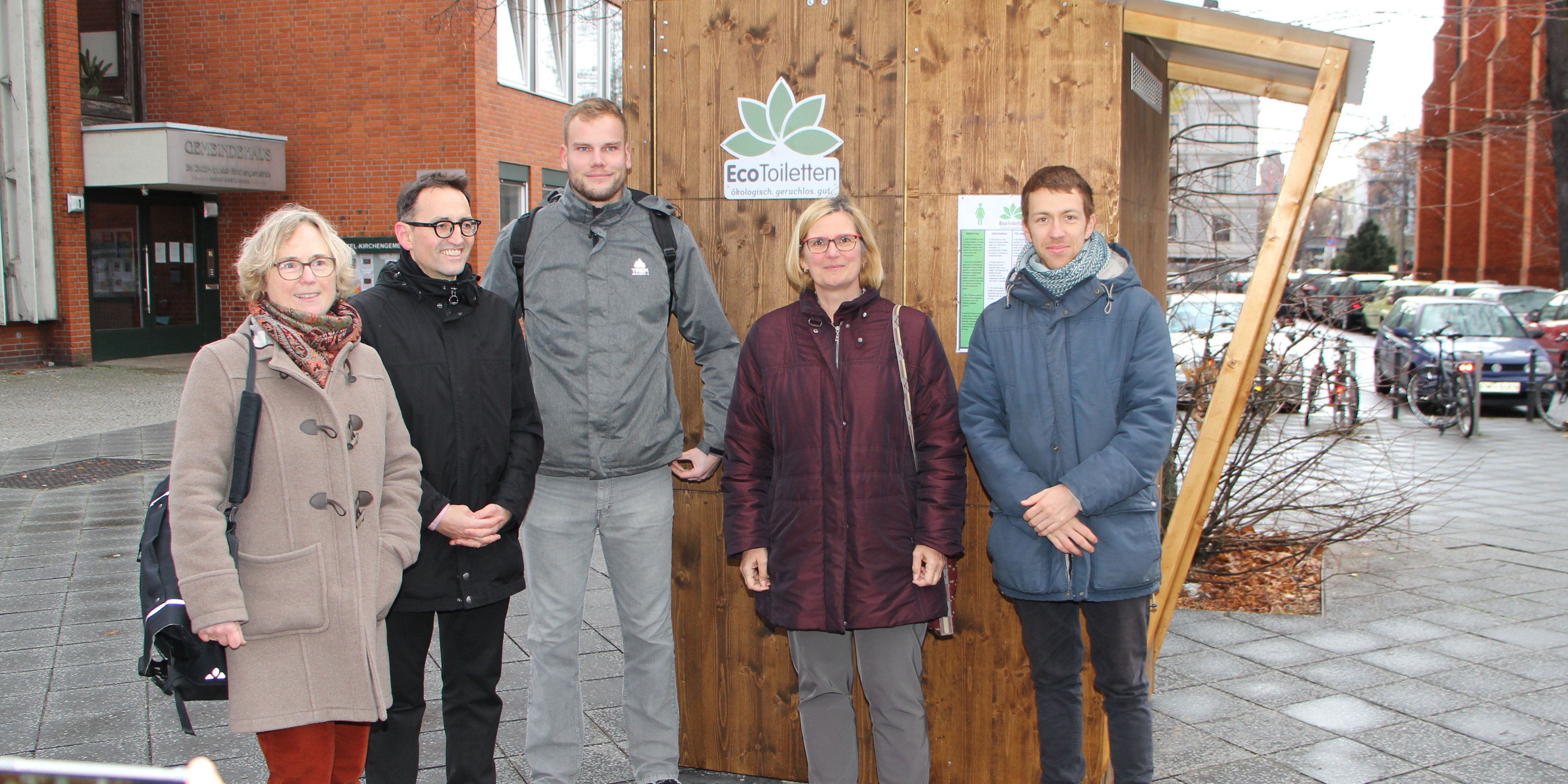 Bildvergrößerung: Die fünf Personen stehen vor dem Toilettenhäuschen aus Holz.