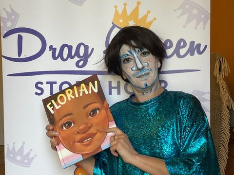 Bildvergrößerung: Eine Person in einer glitzernden Robe und Gesichtsbemalung steht vor einer Wand, auf der Drag Queen Story Hour steht. Ein Buch mit dem Tite Florian wird hochgehalten.