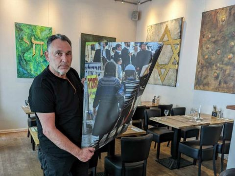 Bildvergrößerung: Ein Mann steht in einem Restaurant und hält ein gekrümmtes und geknicktes Bild in der Hand. Im Hintergrund sind weitere Bilder an den Wänden.