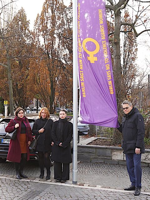 Bildvergrößerung: vier Personen stehen an einem Fahnenmast an der eine lilafarbene Fahne mit dem Symbol für Weiblichlichkeit