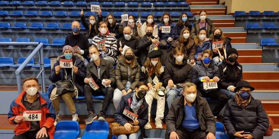 Eine Gruppe Menschen mit Masken sitzt in mehreren Reihen auf einer Tribüne und hält kleine Schilder mit Nummern hoch.