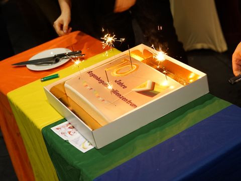 Bildvergrößerung: Auf einer mit Regenbogenflagge gedecktem Tisch steht ein Kuchen mit Wunderkerzen. Auf dem Kuchen steht "10 Jahre Regenbogenfamilienzentrum".