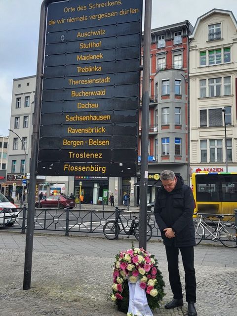 Eine große schwarze Tafel mit Namen von Orten, darunter ein Blumenkranz und ein Mann, der den Kopf im Gedenken gesenkt hat