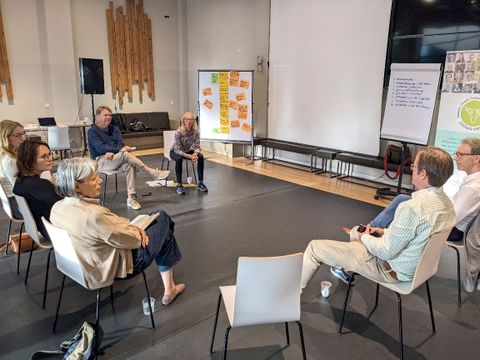 Bildvergrößerung: Sieben Personen sitzen in einem hohen Raum im Halbkreis vor einigen Flipcharts.